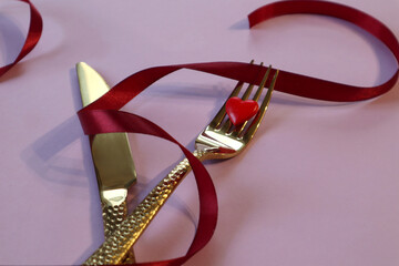 Red heart on a Golden fork, knife, red ribbon, pastel background, close-up, top view-the concept of holding a romantic dinner on holidays