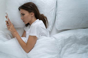Lonely woman with depressed facial expression lying in bed holding mobile smart phone, can't sleep, addicted to social networks, looking at screen and waiting message or call from boyfriend.