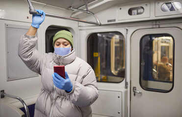 Fototapeta na wymiar Woman in warm clothing and in protective mask and gloves reading online book on the phone while standing on a train