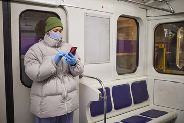 Woman in mask and in warm clothing typing a message on mobile phone while standing on a train in subway