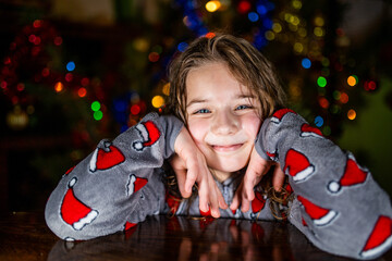 Dziewczynka w świątecznej piżamie uśmiechająca się na tle lampek choinkowych. 