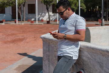 Outdoor photo of a charismatic young student with sunglasses using a smartphone A Latino man...