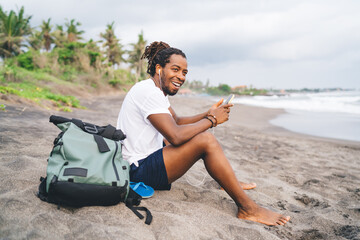 Smiling African American male sitting on seashore with phone