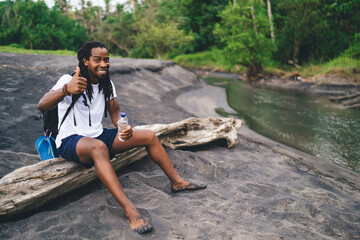 Black man sitting on log in forest with thumb up