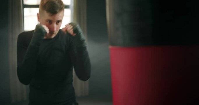 Boxing, athletic man fighter trains his punches, beats a punching bag, training day in the boxing gym, strikes with his feet, back lighting.