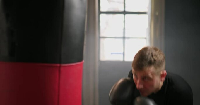 Boxing, athletic man fighter trains his punches, beats a punching bag, training day in the boxing gym, strikes with his feet, back lighting.