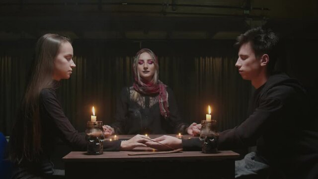 Young couple visits young witch female fortune teller. They holds each ather hands and closes eyes