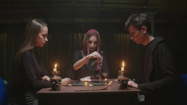 Witch female fortune teller tries to speak with ghost. She uses a blue crystal and board with letters. Two women and one man uses Ouija Board Spirit Game