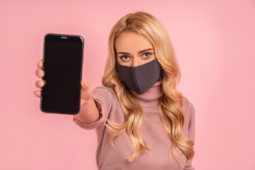 Young blonde girl in pink blouse, sterile face mask isolated on pink background. Epidemic pandemic coronavirus 2019-ncov sars covid-19 flu virus concept. Hold mobile phone with blank empty screen