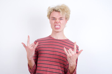 Young handsome Caucasian blond man standing against white background making rock hand gesture and showing tongue