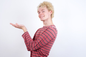 Young handsome Caucasian blond man standing against white background pointing aside with hands open palms showing copy space, presenting advertisement smiling excited happy