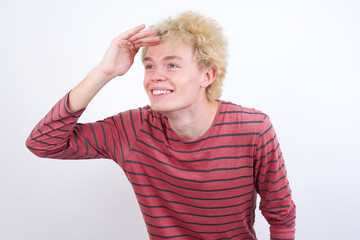 Young handsome Caucasian blond man standing against white background very happy and smiling looking far away with hand over head. Searching concept.