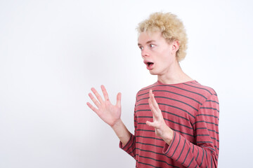 Young handsome Caucasian blond man standing against white background shouts loud, keeps eyes opened and hands tense.