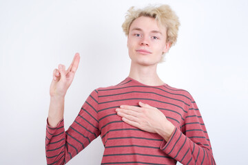 Young handsome Caucasian blond man standing against white background smiling swearing with hand on...