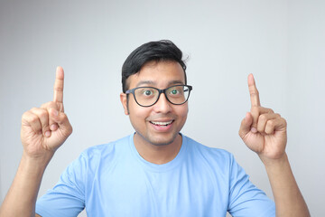 a young indian man in spectacles looking towards the camera and smiling very excited and pointing both fingers up to blank copy space