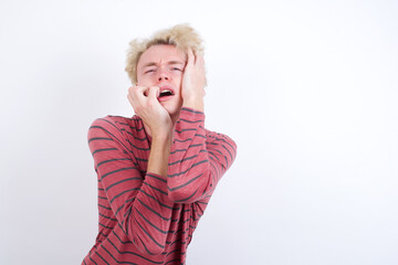 Doleful desperate crying Young handsome Caucasian blond man standing against white background, looks stressfully, frowns face, feels lonely and anxious