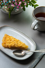 a piece of lemon pie on a plate on a gray table with a bouquet of gypsophila and a mug of tea