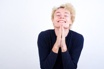 Young handsome Caucasian blond man standing against white background praying with hands together asking for forgiveness smiling confident.