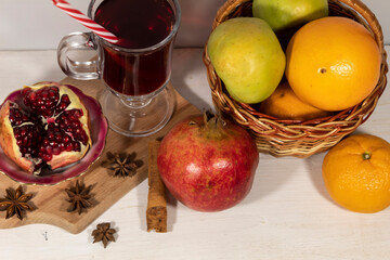 On the table is a glass of pomegranate juice, a basket of apples and citrus fruits. Nearby are pomegranates, cinnamon sticks and star anise.