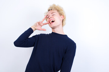 Young handsome Caucasian blond man standing against white background Doing peace symbol with fingers over face, smiling cheerful showing victory