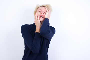 Doleful desperate crying Young handsome Caucasian blond man standing against white background, looks stressfully, frowns face, feels lonely and anxious