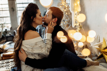 Charming husband gently kiss adorable wife, enjoy tender moment. Handsome man and gorgeous woman sitting on the bed near decorated Christmas tree. Beautiful couple celebrate xmas eve at home