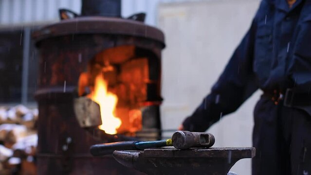Selective focus of anvil and hammer on stool with young female craftswoman standing in front of forge wearing uniform and pocket belt