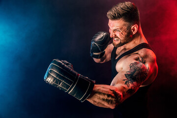 Bearded tattooed sportsman muay thai boxer in black undershirt and boxing gloves fighting on dark...