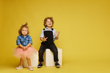 Cute children posing over a yellow background. Sad little girl and happy boy in casual outfits on the yellow background, isolated with copy space