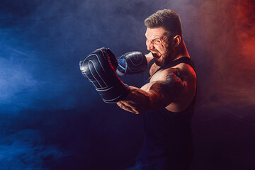 Bearded tattooed sportsman muay thai boxer in black undershirt and boxing gloves fighting on dark...