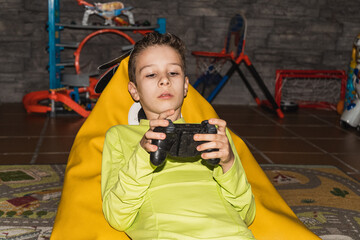 Child playing video games lying on a pouf wearing a yellow T-shirt with the controller in his hand. Lifestyle