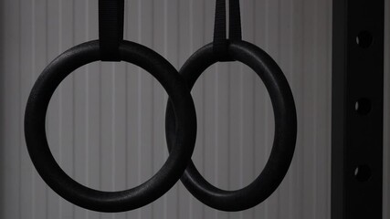 Close-up of set of black gymnastic rings or steady rings hanging and swinging in steel power rack over a white wall. Detail of closed gym with the lights off. Fitness or healthy lifestyle concept
