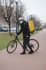 Vertical shot of a courier wearing medical face mask and thermo backpack, walking in the city with his bicycle