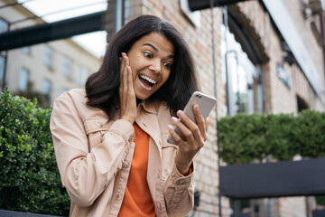 Fototapeta premium Surprised African American woman using mobile phone, looking at digital screen, she winning online lottery, celebration success. Young excited female shopping online with cash back, big sales
