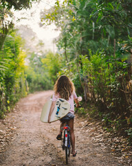 Woman on a bicycle with surfboard on rural road with green trees along. Tropical green. Sport...