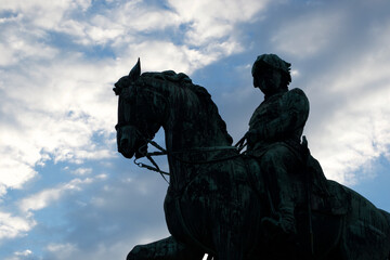 Silhouette of a dark metal statue of a soldier on a horse, standing boldly against a blue, cloudy sky.