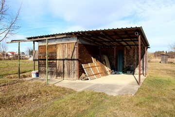 Small old wooden storage shed with open area with wooden construction material and some junk built on concrete foundation surrounded with grass and traditional outdoor wooden toilet in background