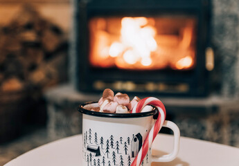 A mug of cocoa or hot chocolate with powdered marshmallows and candy cane by the fireplace