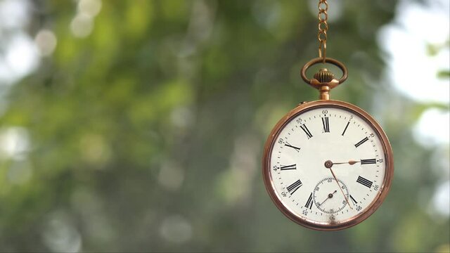 Pocket Watch on Chain Time is Ticking 4K loop features a gold pocket watch with hands spinning around and swinging back and forth with blurred leaves blowing in the wind in the background in a loop