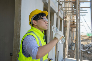Asian male engineer foreman stands to rest and drinks in a bottle at a home construction project.