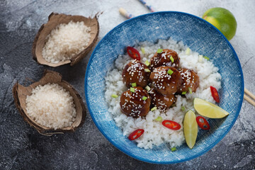 Blue bowl with asian-style white rice and teriyaki sauce meatballs, elevated view on a dark grey stone background, studio shot