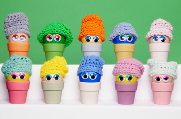 Colourful Easter eggs, made of recycled plastic, with wobbly eyes and colourful knitted hats, aligned horizontally and sitting in  bamboo egg holders against an apple green background. 