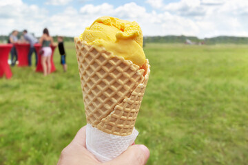 Ice cream in a waffle cone, in your hand. Holiday fair in the open air, unfocused unrecognizable people in the distance