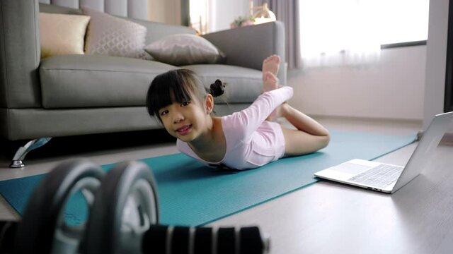 Little asian girl doing yoga plank and watching online lessons on laptop, exercising in the kitchen. A child plays sports during the COVID-19 pandemic.