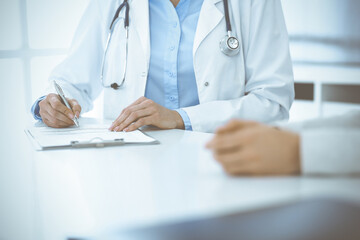 Fototapeta premium Unknown woman-doctor and female patient sitting and talking at medical examination in clinic, close-up. Therapist wearing blue blouse is filling up medication history record. Medicine concept