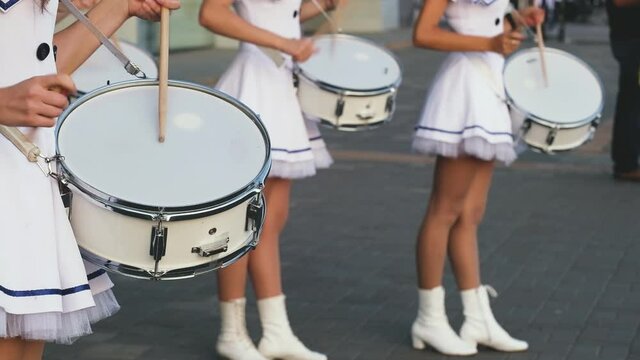 Young women  in white marine short dresses play drums on a parade on summer day. Girls musicians drummers from municipal orchestra performing on festival. Close-up of hands with wooden sticks.