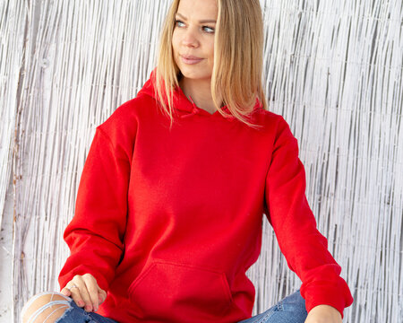 Smiling blond woman wearing red blouse or hoodie sits in front of camera. On her hoodie in place for christmas inscription. Winter outfit with copy space for mockup.