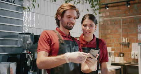 Attractive couple of barista workers communicating using smartphone social media application inside modern cafe interior. Coffee store. - Powered by Adobe