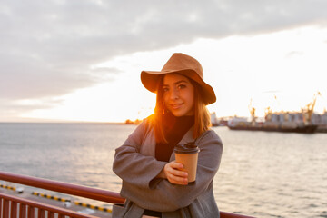 Stylish woman in felt hat and coat drinks coffee on the go at sunrise at sea