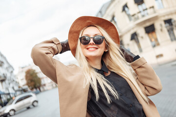 Portrait of young stylish blonde woman in autumn coat, sunglasses, gloves and felt hat in European...